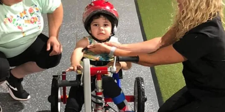 An adult adjusts equipment while a young rider sits on a red adaptive Tryke